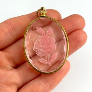 May include: A vintage oval-shaped pendant with a delicate pink rose carved into a clear glass cameo. The pendant is set in a gold-toned metal frame.