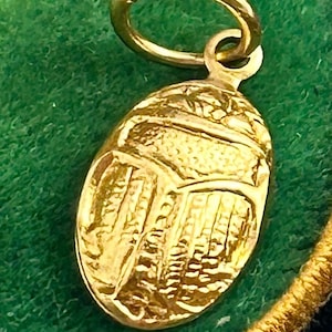 May include: A gold-colored scarab beetle charm with a loop for attaching to a necklace or bracelet. The charm is highly detailed, with etched lines and patterns across its surface. The background is a dark green fabric.