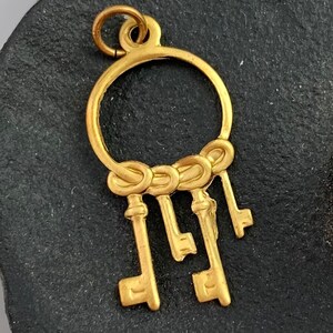 Op de afbeelding: Gouden bedel met een cirkel waar vijf sleutels aan hangen. De sleutels zijn allemaal in een vergelijkbare stijl met een ronde bovenkant en een puntig uiteinde.
