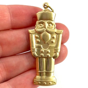 May include: A gold-colored metal nutcracker ornament. The nutcracker is wearing a hat and has a mustache and beard. The ornament is detailed with small dots and lines.