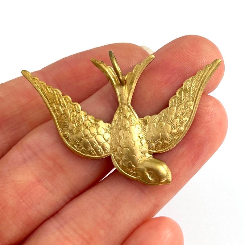 May include: A gold-toned metal pendant in the shape of a stylized bird with its wings spread. The bird has a detailed design with textured feathers.