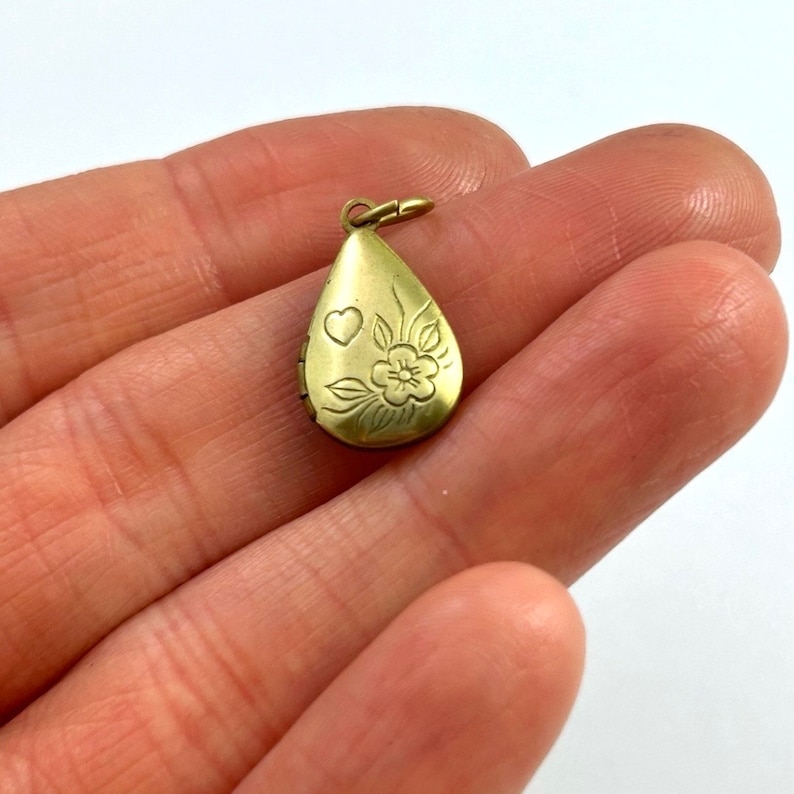 May include: Gold teardrop-shaped locket with an engraved floral design and a heart. The locket has a hinged opening.
