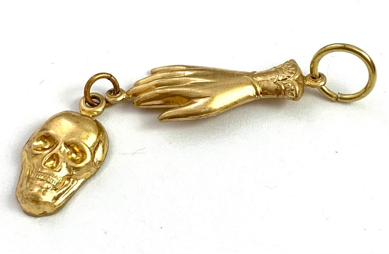 Op de afbeelding: Een gouden bedel met een gedetailleerde schedel en een hand met een manchet. De bedel is bevestigd aan een gouden ring.