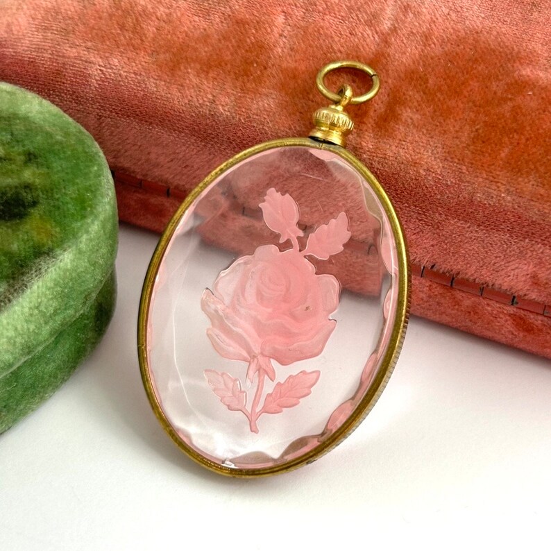 May include: A gold-toned pendant with a clear glass cameo featuring a pink rose etched into the glass.