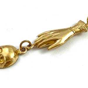 Op de afbeelding: Een gouden hanger met een gedetailleerde schedel en een uitgestrekte hand. De hand is versierd met een decoratieve manchet aan de pols.