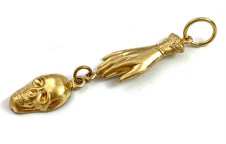 Op de afbeelding: Een gouden hanger met een gedetailleerde schedel en een uitgestrekte hand. De hand is versierd met een decoratieve manchet aan de pols.