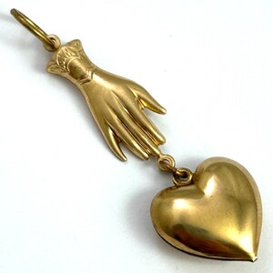 May include: A gold pendant featuring a hand holding a heart. The hand is detailed with fingers and a wrist. The heart is a solid, rounded shape.