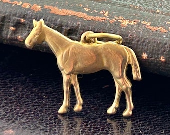 Puede incluir: Un colgante de caballo de color dorado. El caballo está de pie con la cabeza girada hacia la izquierda. El colgante está hecho de metal y tiene un acabado brillante.
