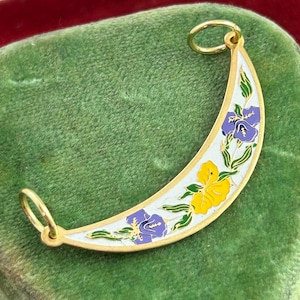 May include: Gold-toned crescent pendant with a white enamel base, adorned with purple and yellow floral designs. The pendant has a loop at each end for attaching to a chain. The floral details are outlined in green.