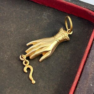K&ouml;nnte beinhalten: Ein goldfarbener Anh&auml;nger in Form einer Hand, die ein Fragezeichen h&auml;lt. Die Hand ist mit komplizierten Schnitzereien verziert und das Fragezeichen ist mit einer kleinen Kette am Handgelenk befestigt.