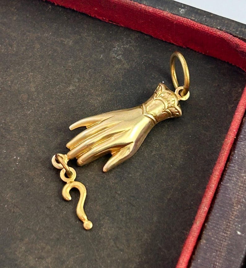K&ouml;nnte beinhalten: Ein goldfarbener Anh&auml;nger in Form einer Hand, die ein Fragezeichen h&auml;lt. Die Hand ist mit komplizierten Schnitzereien verziert und das Fragezeichen ist mit einer kleinen Kette am Handgelenk befestigt.