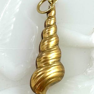 May include: A gold-colored seashell charm with a spiral design. The charm has a loop at the top for attaching to a necklace or bracelet. The seashell is detailed with ridges and curves.