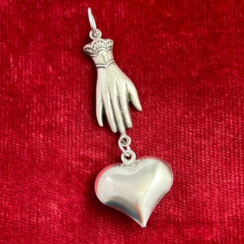 Puede incluir: Un colgante de plata con forma de mano sosteniendo un coraz&oacute;n. La mano est&aacute; detallada con un brazalete y el coraz&oacute;n es liso y redondeado.