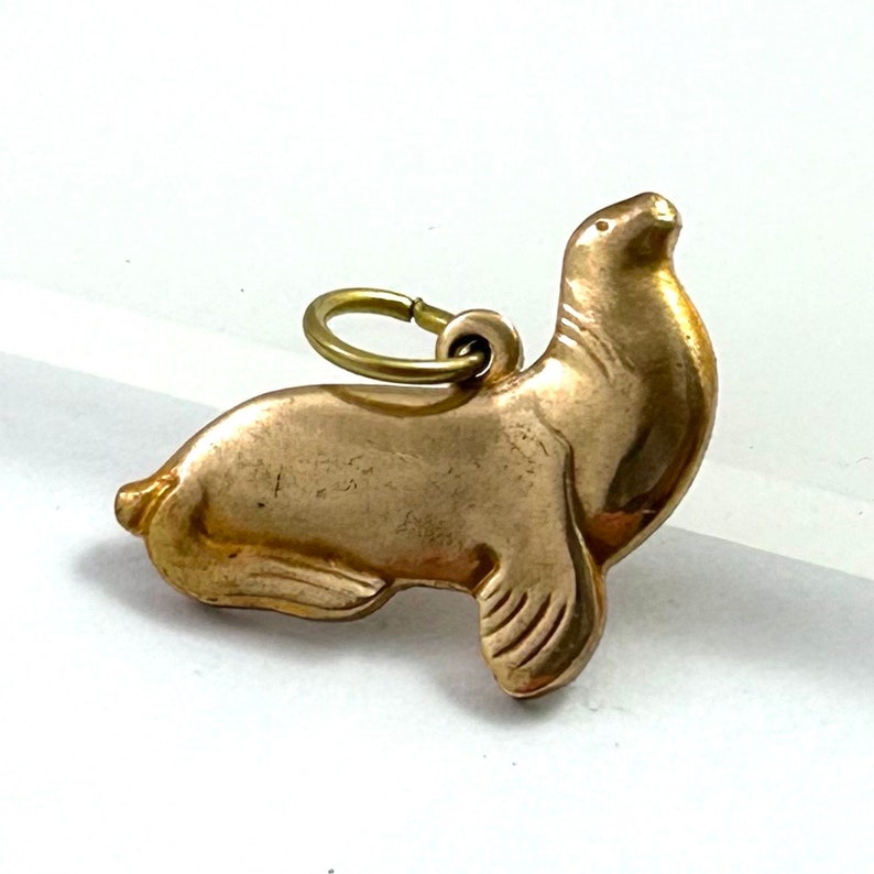 May include: A gold-colored charm in the shape of a seal. The charm has a loop at the top for attaching to a necklace or bracelet.