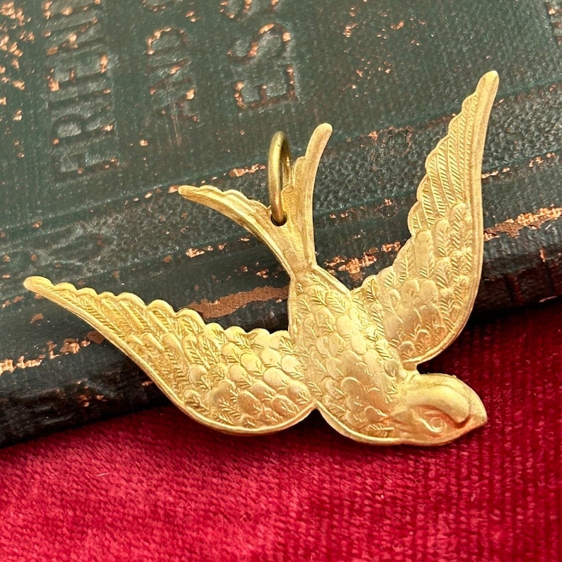 May include: A gold-toned metal bird pendant with detailed etched feathers. The bird is in flight with its wings spread wide.