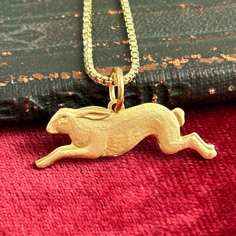 May include: A gold-coloured necklace with a detailed rabbit pendant. The rabbit is depicted in a running pose with its ears raised.