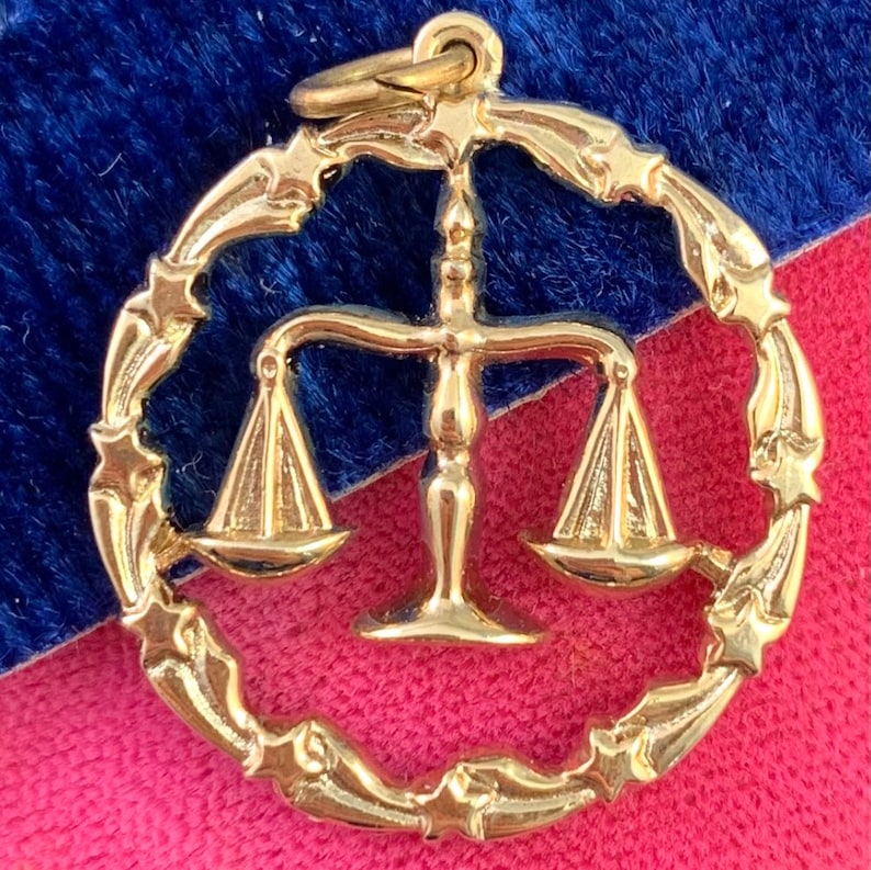 May include: Gold-toned zodiac charm featuring a stylized depiction of the scales of Libra, surrounded by a circle of stars.