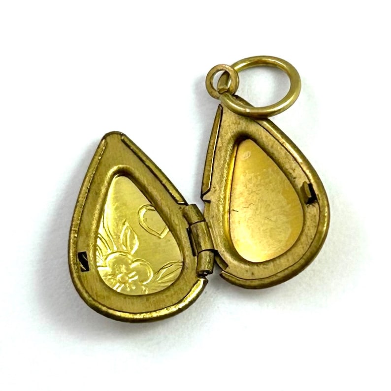 May include: A gold-toned teardrop-shaped locket with a floral design on the front and a plain back. The locket is open and has a hinge on the side. It has a small ring at the top for attaching a chain.