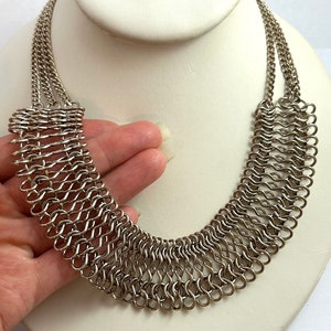 May include: A silver chainmail necklace with a wide, curved design. The necklace is made of small, interconnected metal rings.