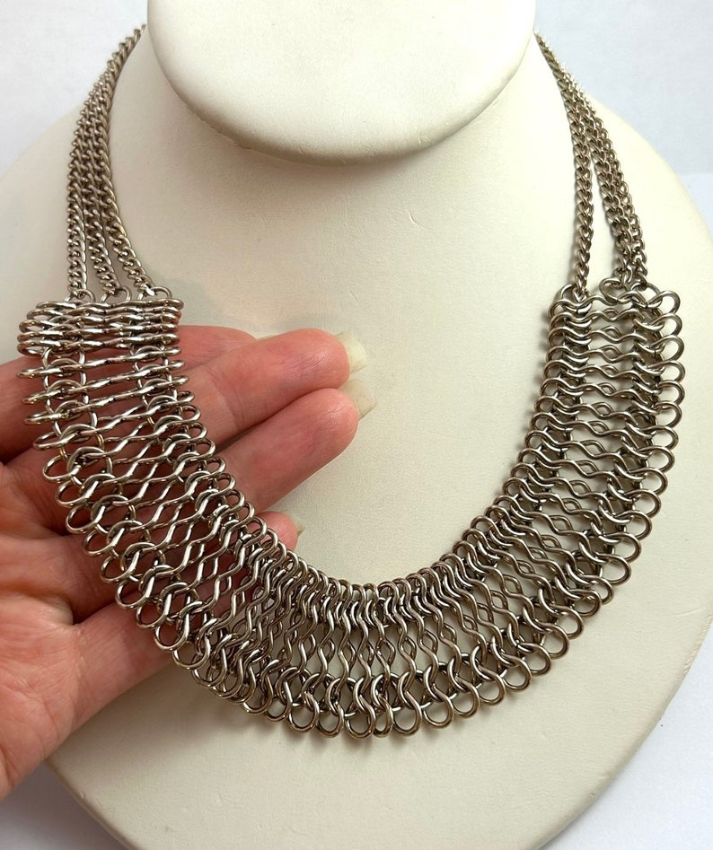 May include: A silver chainmail necklace with a wide, curved design. The necklace is made of small, interconnected metal rings.