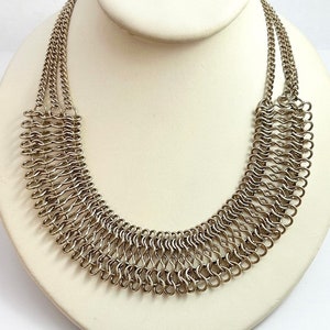 May include: A silver chainmail necklace with a wide, curved design. The necklace is made of many small, interconnected metal rings.
