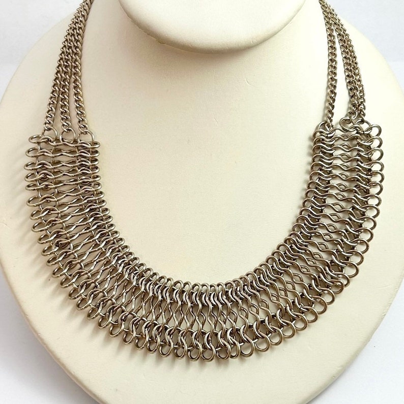 May include: A silver chainmail necklace with a wide, curved design. The necklace is made of many small, interconnected metal rings.