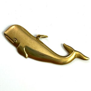May include: A gold-coloured brass whale charm pendant. The whale is depicted in a side profile with its tail fin extended to the right. The charm has a small loop at the top for attaching to a necklace or bracelet.