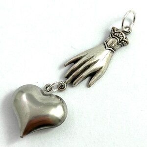 Puede incluir: Collar con dije de plata que representa una mano que se extiende hacia un coraz&oacute;n. La mano est&aacute; detallada con un brazalete y el coraz&oacute;n es liso y redondo.
