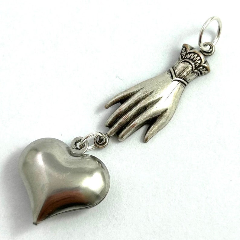 Puede incluir: Collar con dije de plata que representa una mano que se extiende hacia un coraz&oacute;n. La mano est&aacute; detallada con un brazalete y el coraz&oacute;n es liso y redondo.