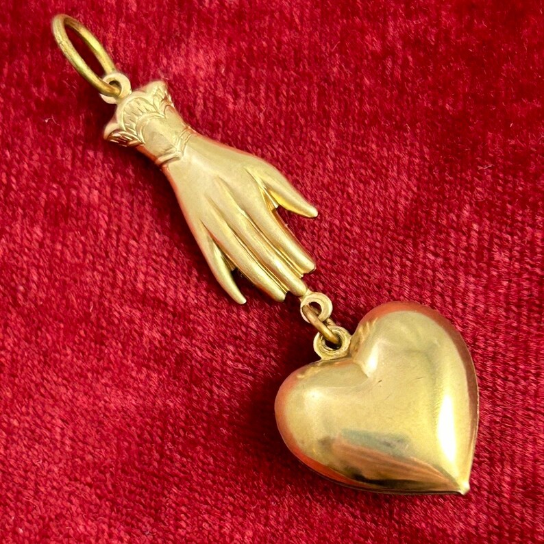 May include: A gold pendant featuring a hand holding a heart. The hand is detailed with a cuff and fingers. The heart is solid and has a smooth surface.