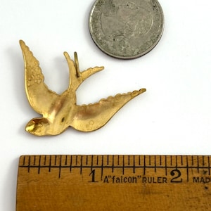 May include: A gold-colored metal bird pendant in the shape of a swallow. The pendant is about 1 inch long and is shown next to a US quarter for size comparison.