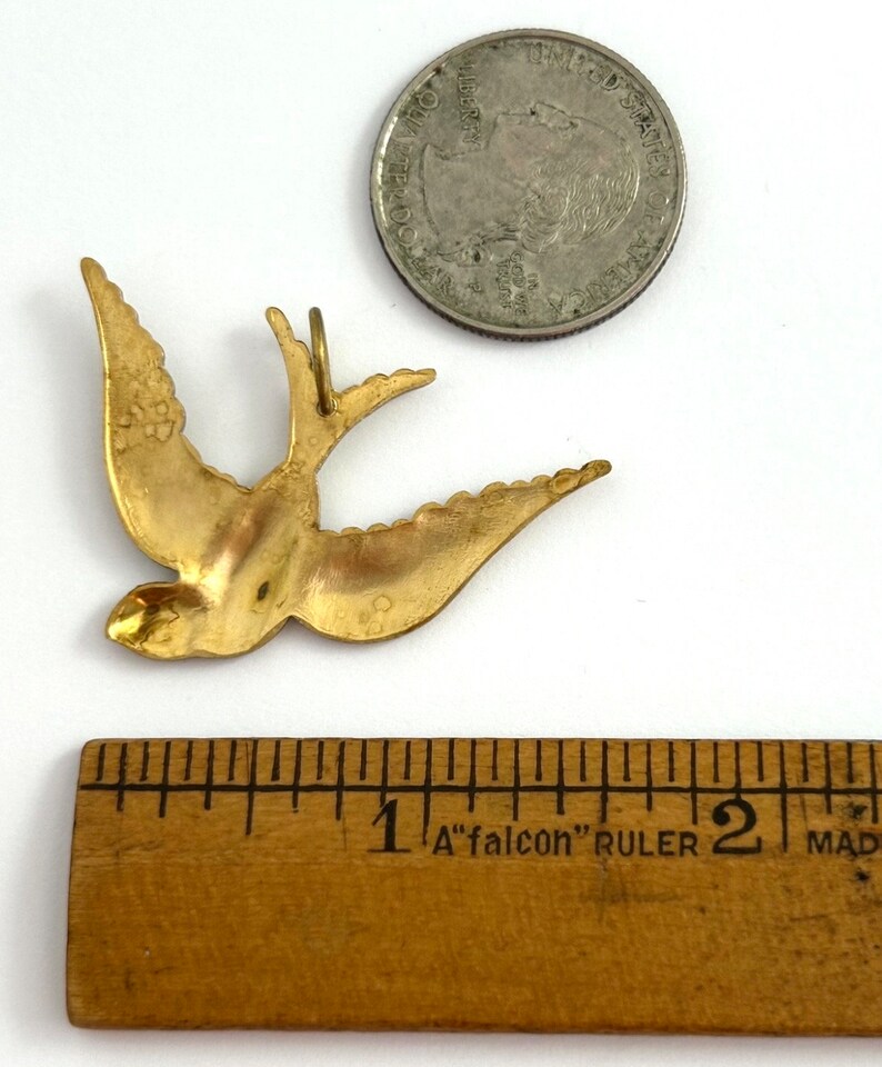 May include: A gold-colored metal bird pendant in the shape of a swallow. The pendant is about 1 inch long and is shown next to a US quarter for size comparison.