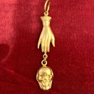 Op de afbeelding: Een gouden hanger met een hand met een schedelbedel eraan. De hanger is op een rode fluwelen achtergrond.