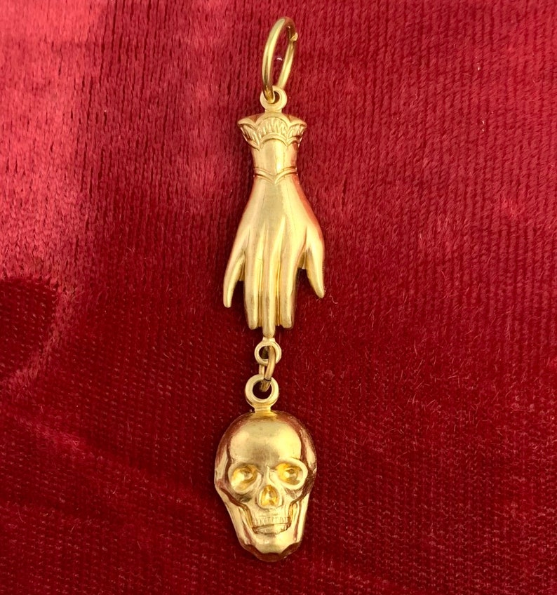 Op de afbeelding: Een gouden hanger met een hand met een schedelbedel eraan. De hanger is op een rode fluwelen achtergrond.