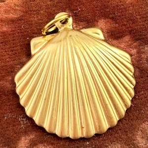 May include: A gold-colored seashell pendant with a ring at the top for hanging. The pendant has a detailed, ridged design, and the overall shape is a scallop shell. The background is a textured, reddish-brown surface.