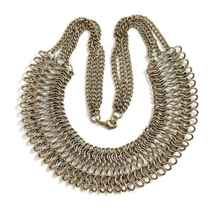 May include: A gold-toned metal chain necklace with a large, intricate, woven design. The necklace is a statement piece and has a clasp closure.