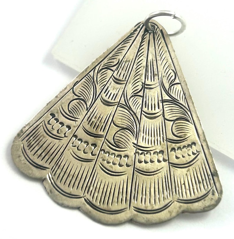 Puede incluir: Colgante plateado con dise&ntilde;o en forma de abanico. El colgante presenta intrincados patrones grabados, incluyendo l&iacute;neas curvas y peque&ntilde;as formas repetitivas. Un peque&ntilde;o anillo est&aacute; unido en la parte superior para colgarlo. El colgante tiene una apariencia ligeramente envejecida.
