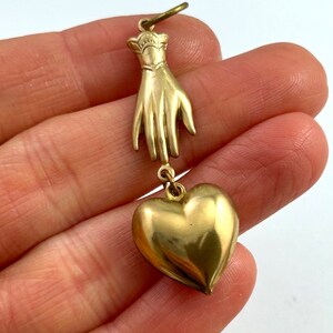 May include: A gold charm pendant featuring a hand with a heart charm hanging from it.