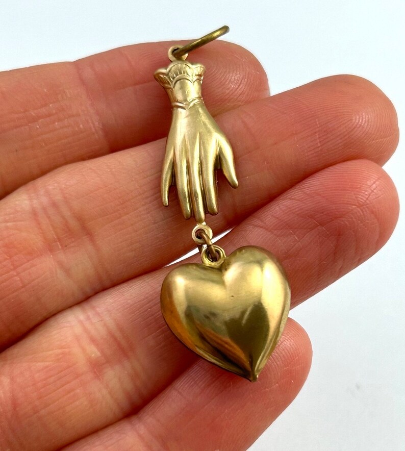 May include: A gold charm pendant featuring a hand with a heart charm hanging from it.
