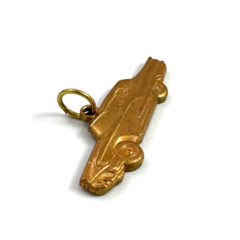 Puede incluir: Un amuleto de metal dorado en forma de coche antiguo. El amuleto tiene un bucle en la parte superior para sujetarlo a un collar o pulsera.