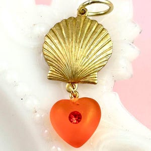 May include: A gold-colored seashell charm with a dangling orange heart pendant. The heart features a red gemstone in the center. The charm is suspended from a gold ring.