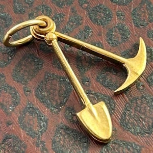 May include: A gold-colored charm in the shape of a shovel and pickaxe. The charm has a loop at the top for attaching to a necklace or bracelet.