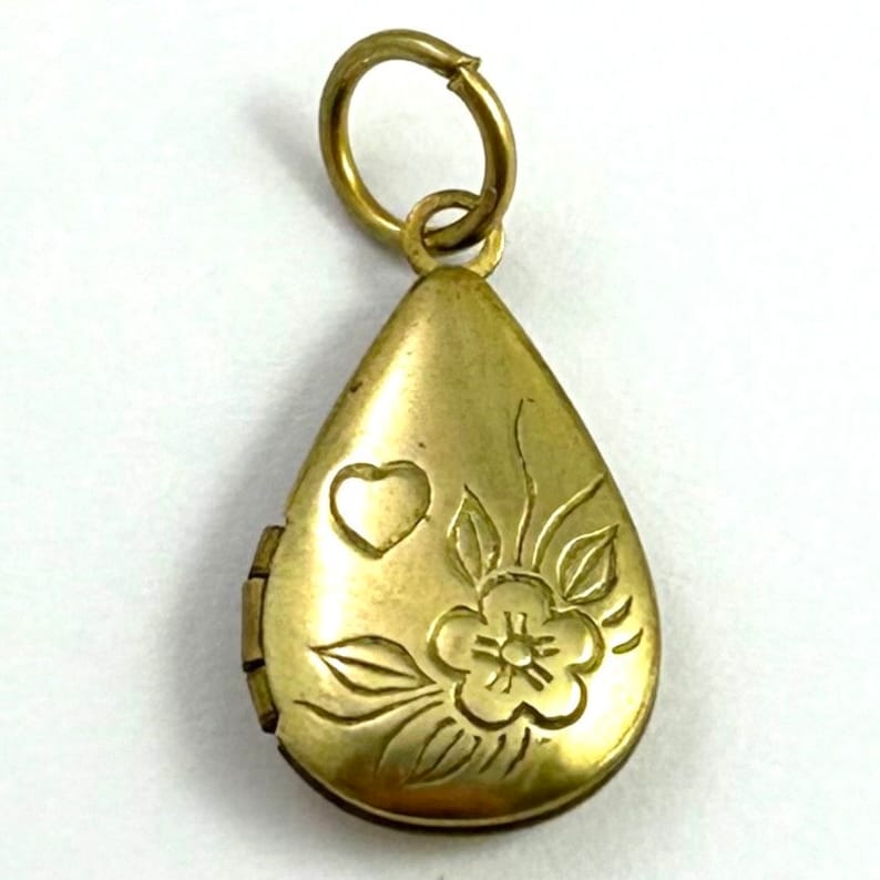 May include: A gold-toned teardrop-shaped locket with a floral design and a heart. The locket has a hinged opening and a loop for attaching a chain.