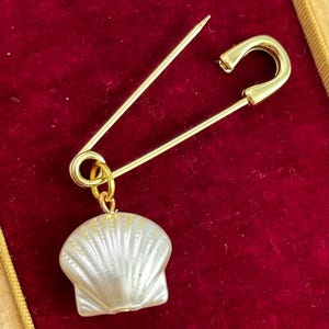 May include: A gold-colored safety pin with a pearlescent white seashell charm. The pin is set against a red velvet background, creating a striking contrast. The seashell charm has detailed ridges.