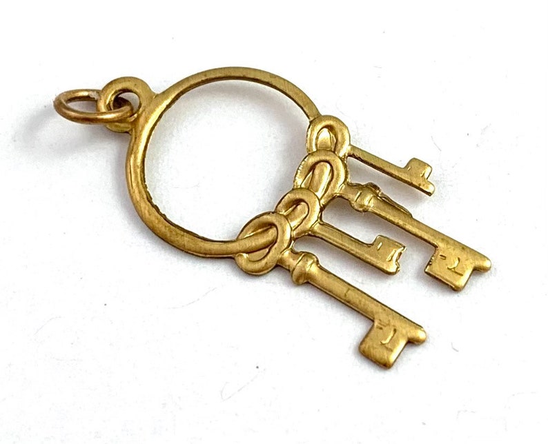 Op de afbeelding: Gouden bedel met een cirkel waaraan vier sleutels hangen. De sleutels zijn met een knoop aan elkaar gebonden.