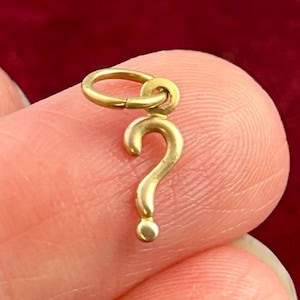 May include: A small, gold-colored question mark charm with a circular loop at the top for attachment. The charm is a simple, elegant design, likely for a necklace or bracelet. The background is a blurred, dark red color.