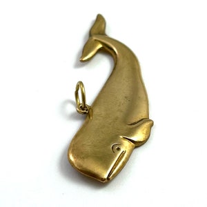 May include: A gold-coloured whale charm pendant. The whale is facing to the right and has a loop at the top for attaching to a necklace or bracelet.