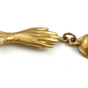 Op de afbeelding: Een gouden bedel met een hand en een schedel. De hand is open en de schedel kijkt naar rechts. De bedel is bevestigd aan een ring.