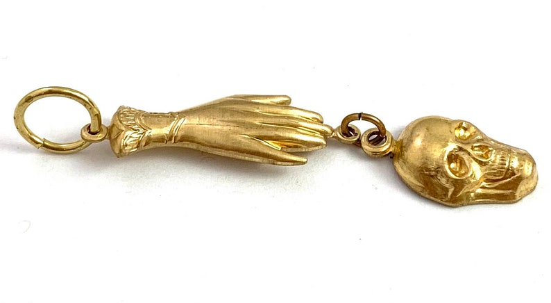 Op de afbeelding: Een gouden bedel met een hand en een schedel. De hand is open en de schedel kijkt naar rechts. De bedel is bevestigd aan een ring.