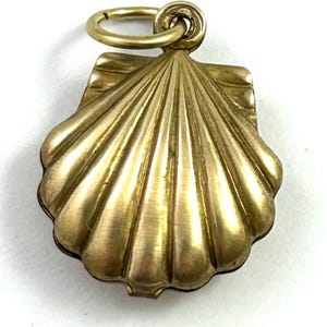 May include: A gold-colored seashell pendant with a textured surface and a small loop for a chain. The pendant has a detailed, fan-like design, and is likely made of brass or a similar metal. The pendant is a jewelry item.